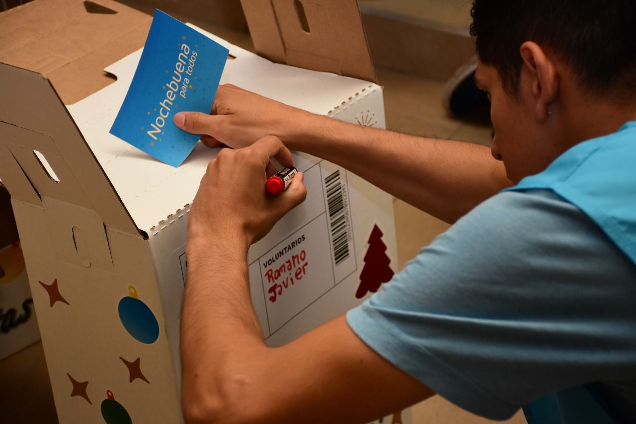 Un voluntario prepara una caja decorada para una colecta navideña, sosteniendo un cartel que dice "Nochebuena para todos", promoviendo la solidaridad comunitaria.