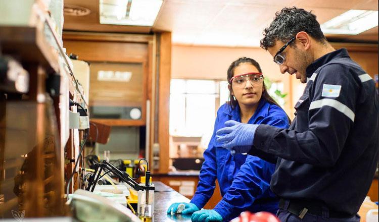 Profesionales colaboran en prácticas laborales en un laboratorio, usando equipo de seguridad y discutiendo procedimientos técnicos.