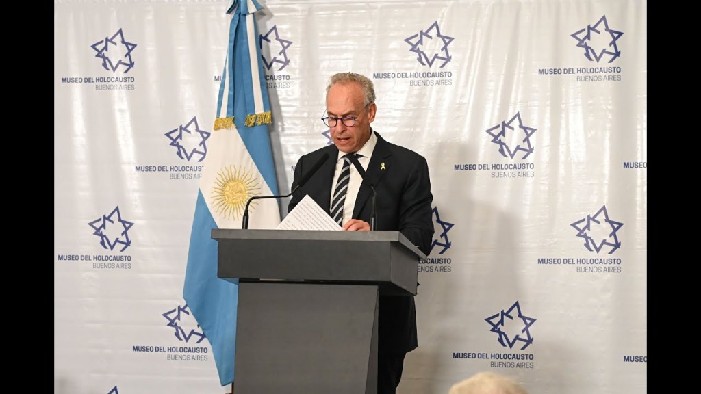 El Museo del Holocausto de Buenos Aires recordó la Shoá en un evento con un orador frente a un atril, acompañado por la bandera argentina y el logo institucional.